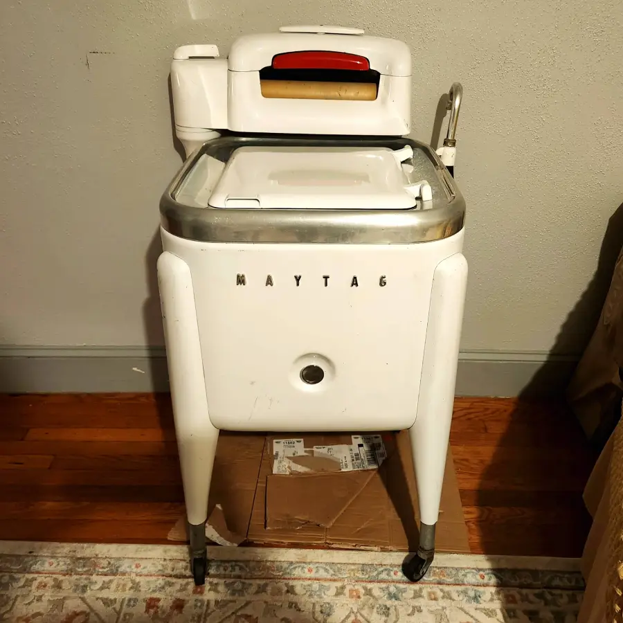 Vintage white Maytag wringer washing machine with a roller at the top, standing on wheels on a hardwood floor near a wall and carpet. A cardboard box is placed underneath the machine.