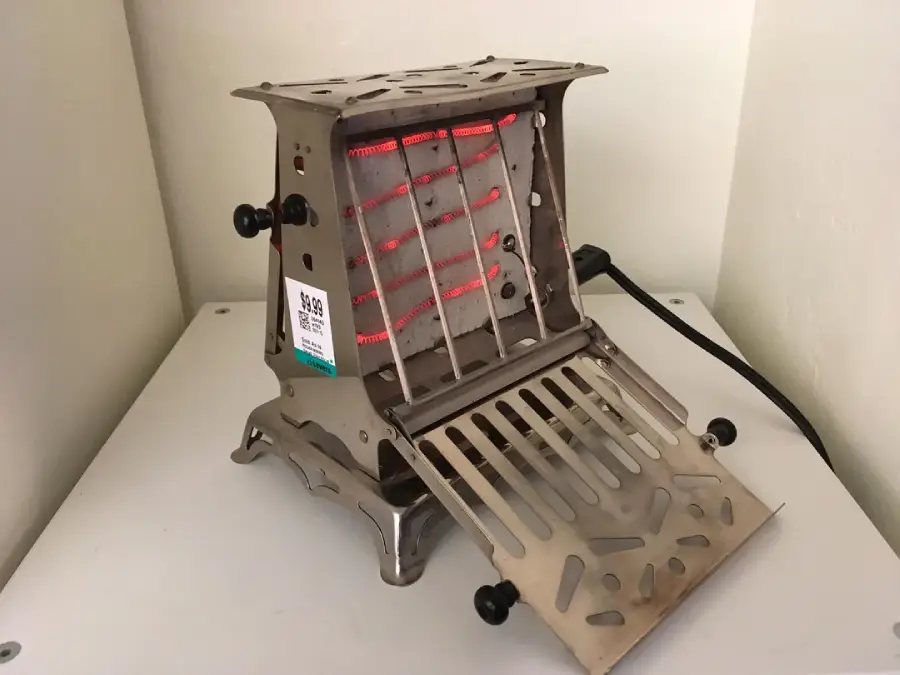 A small metal toaster with an open front panel exposing heated coils, placed on a white surface in the corner of a room.
