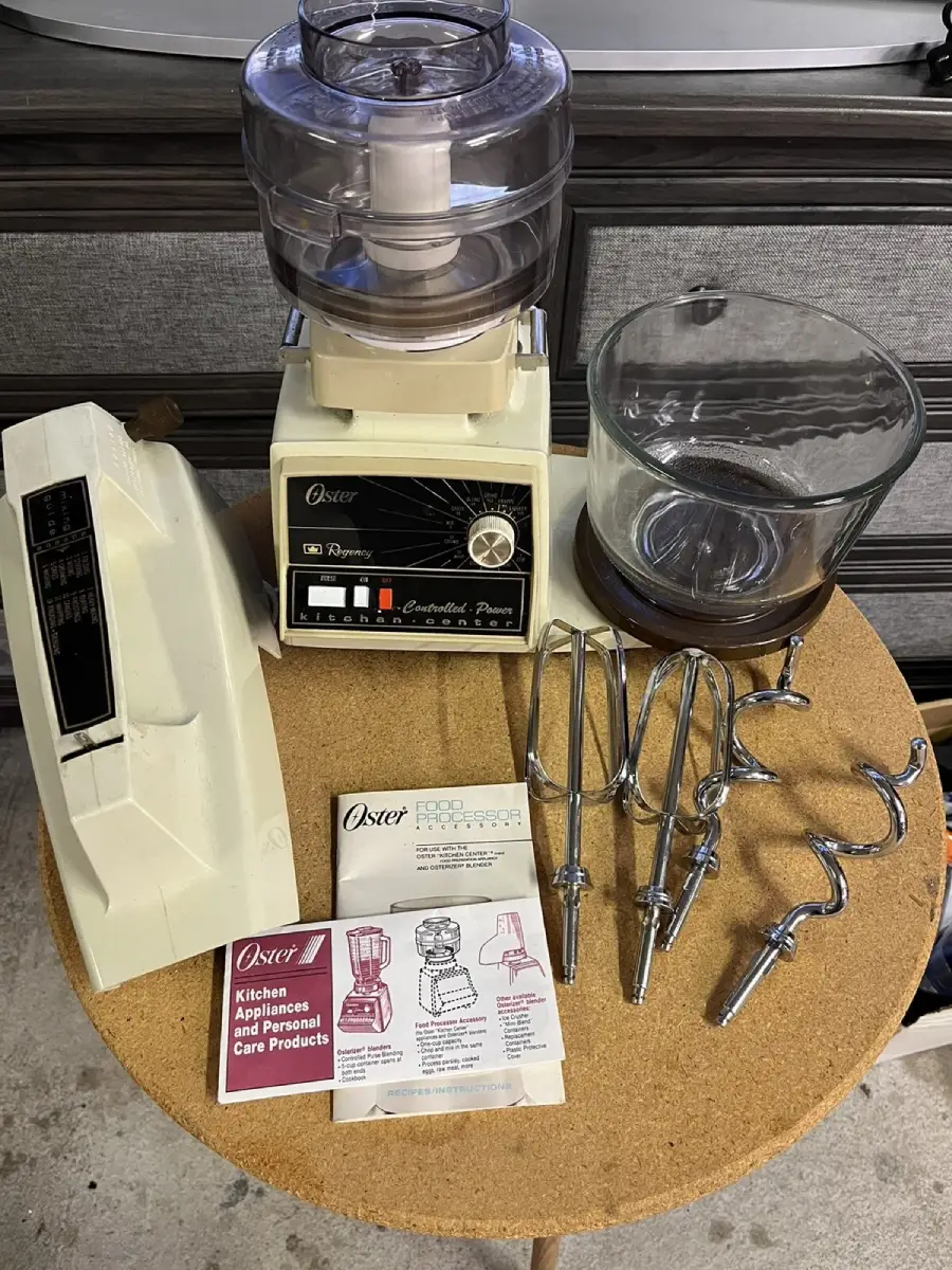 A vintage Oster food processor and mixer set with attachments, manuals, and a glass bowl displayed on a round wooden table. Mixer attachments include beaters and dough hooks.