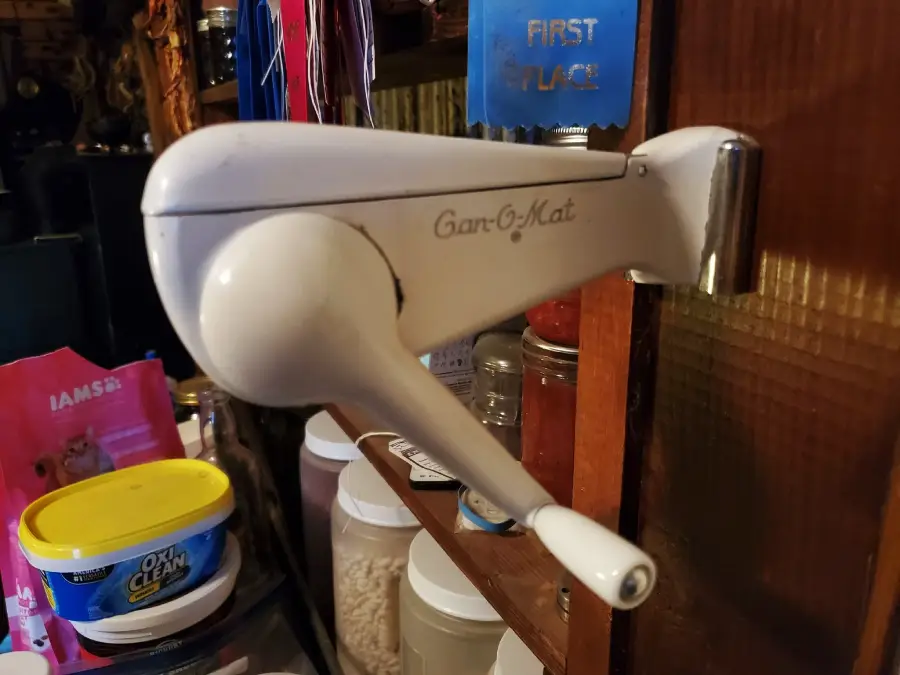 Wall-mounted vintage can opener labeled “Can-O-Mat” with a crank handle, attached to a wooden surface. Nearby are pet food containers, jars, and household items in a kitchen or pantry setting.