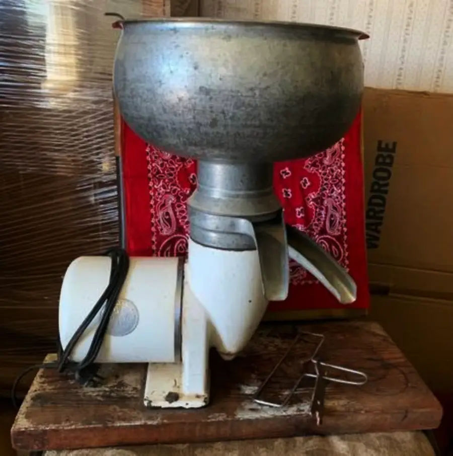 A vintage metal cream separator sits on a wooden table, with a large bowl on top and spouts for separating milk and cream. Red cloth and cardboard boxes are in the background.