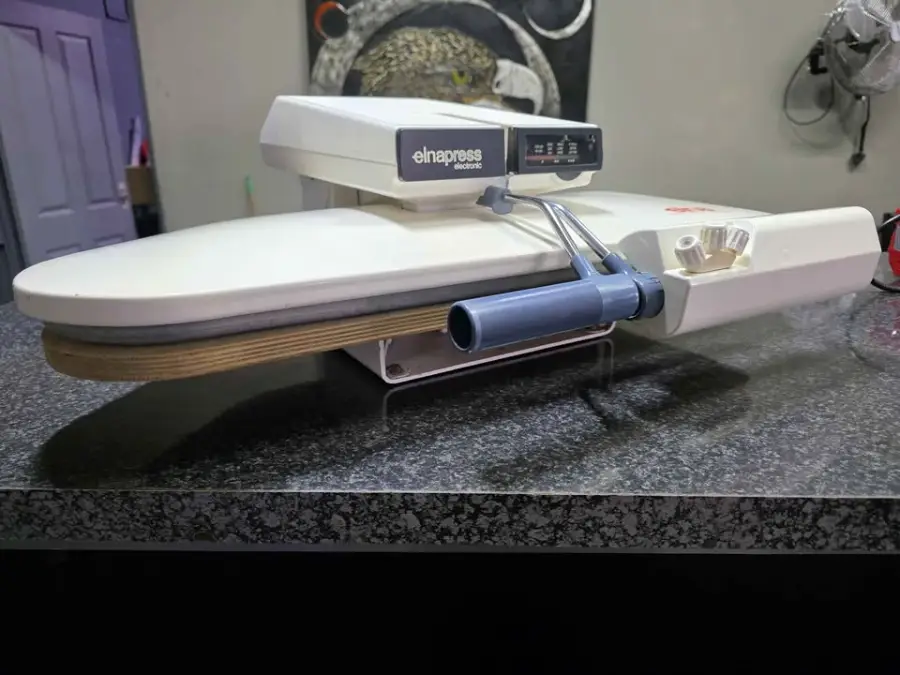 A white ElnaPress ironing press sits on a countertop, with controls and handle visible. Background includes a wall, door, and some hanging items.