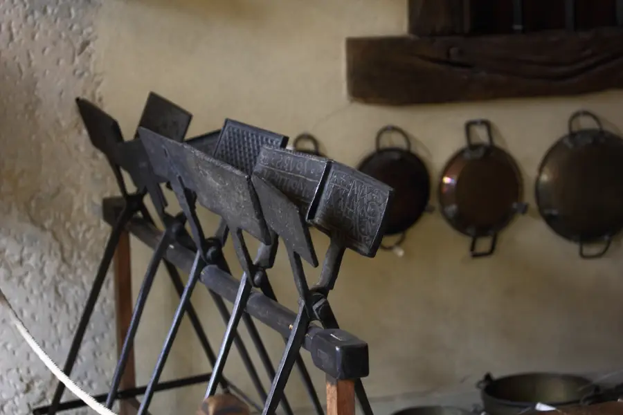 Several vintage cast iron waffle irons rest on a rack in an old-fashioned kitchen, with pots and pans hanging on the wall in the background.