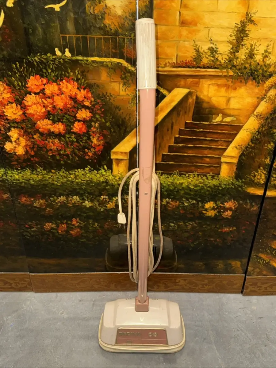 A vintage upright vacuum cleaner with a pink handle and coiled cord stands on a gray floor, positioned in front of a painted backdrop featuring stairs, greenery, and a yellow brick wall.
