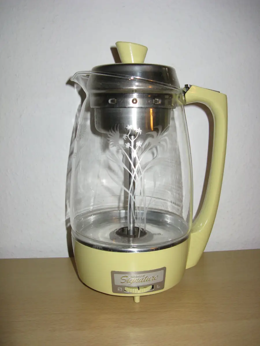 A vintage electric coffee percolator with a clear glass body, floral design, beige handle, and matching base, sitting on a light wooden surface against a plain white wall.