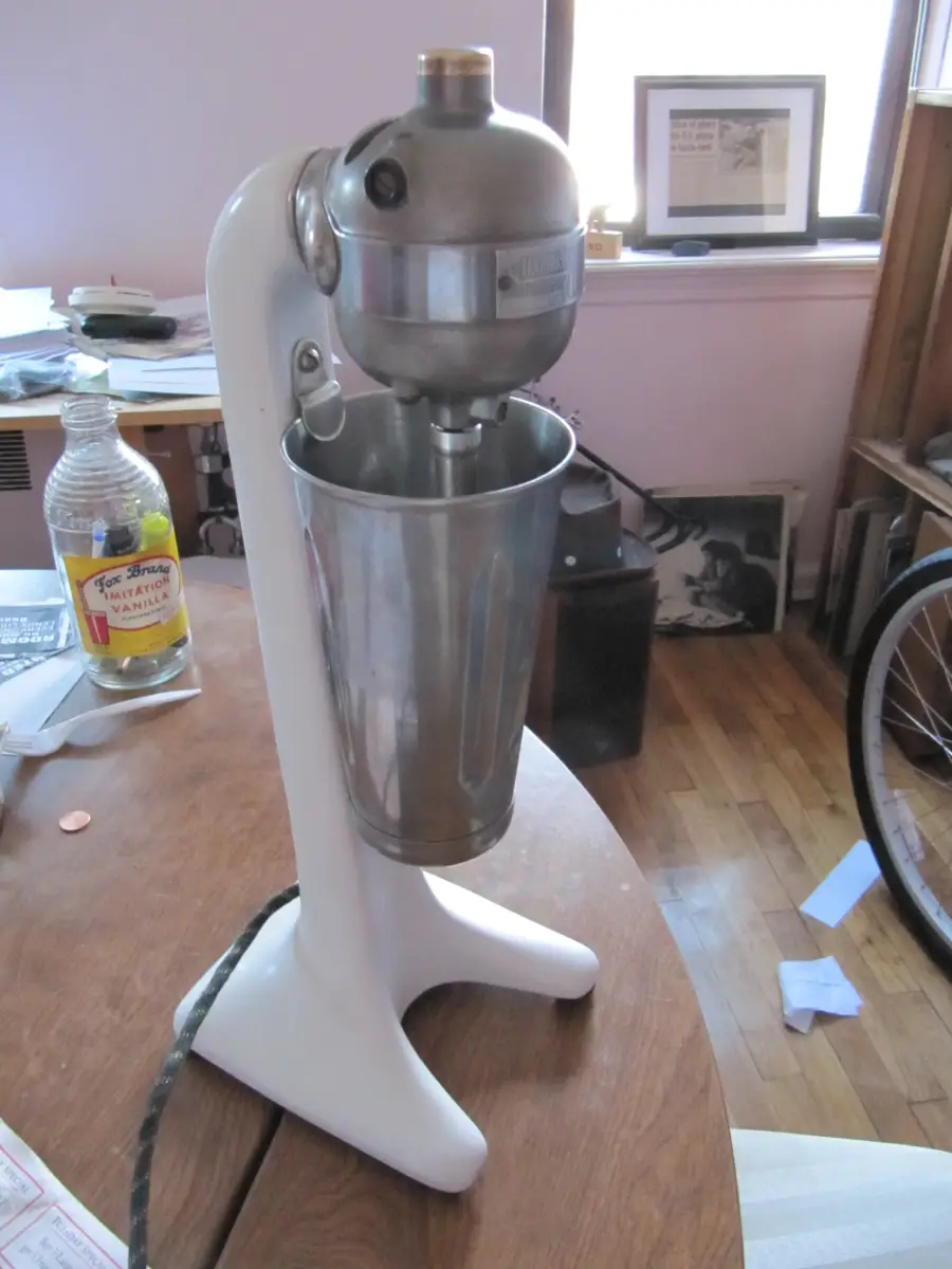 A vintage white and metal milkshake mixer sits on a wooden table in a cluttered room with papers, a bike wheel, and a window in the background.