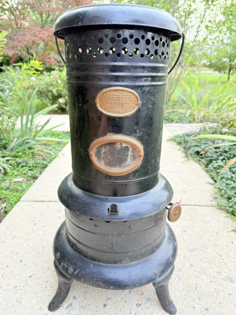 A vintage black cast iron heater or stove with a round base, glass window, and metallic label, standing on a sidewalk with greenery in the background.