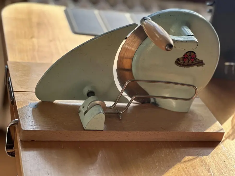 A vintage hand-crank food slicer with a metal blade, wooden base, and wooden handle is sitting on a table in sunlight.