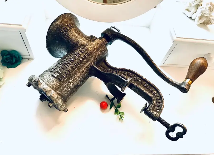 A vintage metal meat grinder with a wooden handle lies on a white surface, surrounded by small decorative items, including a red bead and green leaves.