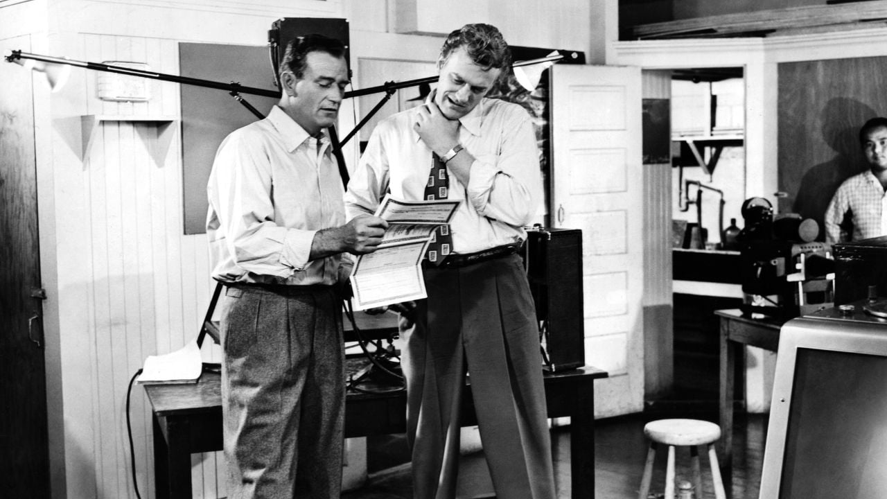 Two men in dress shirts and slacks stand in an office, looking at documents together. One smiles while holding papers. There is office equipment, a stool, and another man in the background. The photo is in black and white.