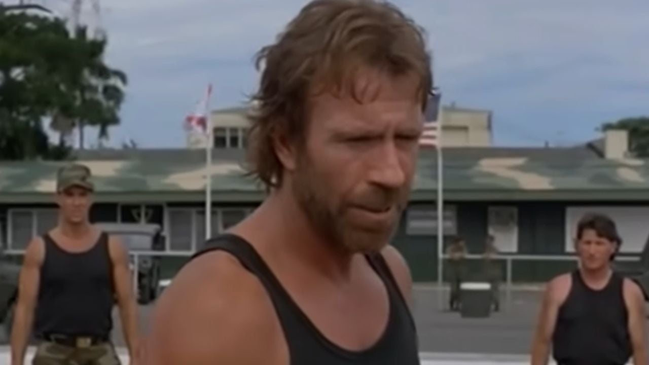 A man with a rugged beard in a black tank top stands outdoors, looking serious. Two men in camouflage clothing stand behind him. A building and trees are visible in the background.