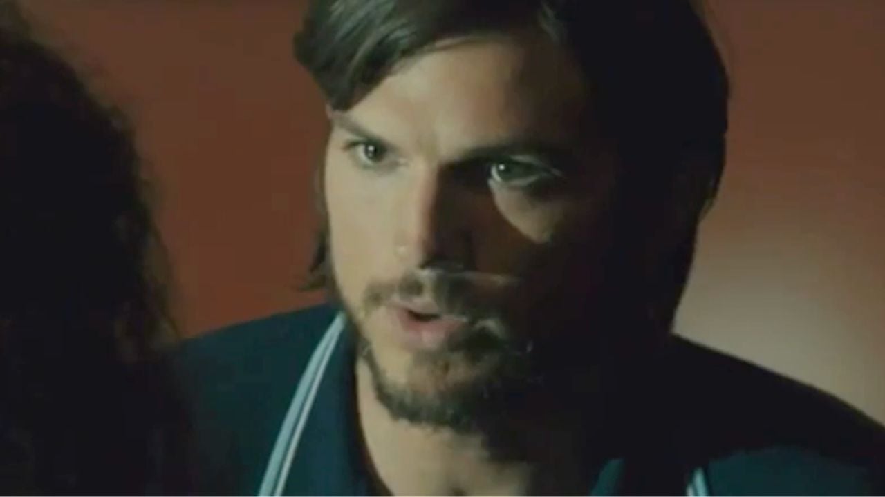 A man with medium-length brown hair, a beard, and mustache looks intently at someone off-camera. The background is softly lit with warm tones, and the man appears serious and engaged in conversation.