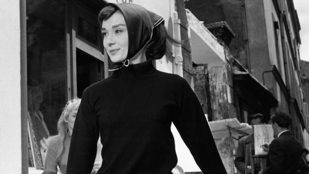 A woman in a dark turtleneck and headscarf stands on a city street, looking to the side. The background shows buildings and people. The image is black and white.