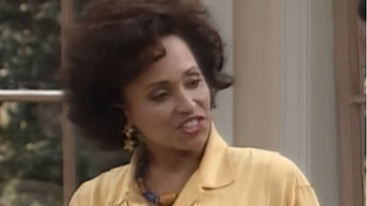 A woman with curly dark hair wearing a yellow blouse and gold earrings stands indoors, speaking and smiling slightly. A window with greenery outside is visible in the background.