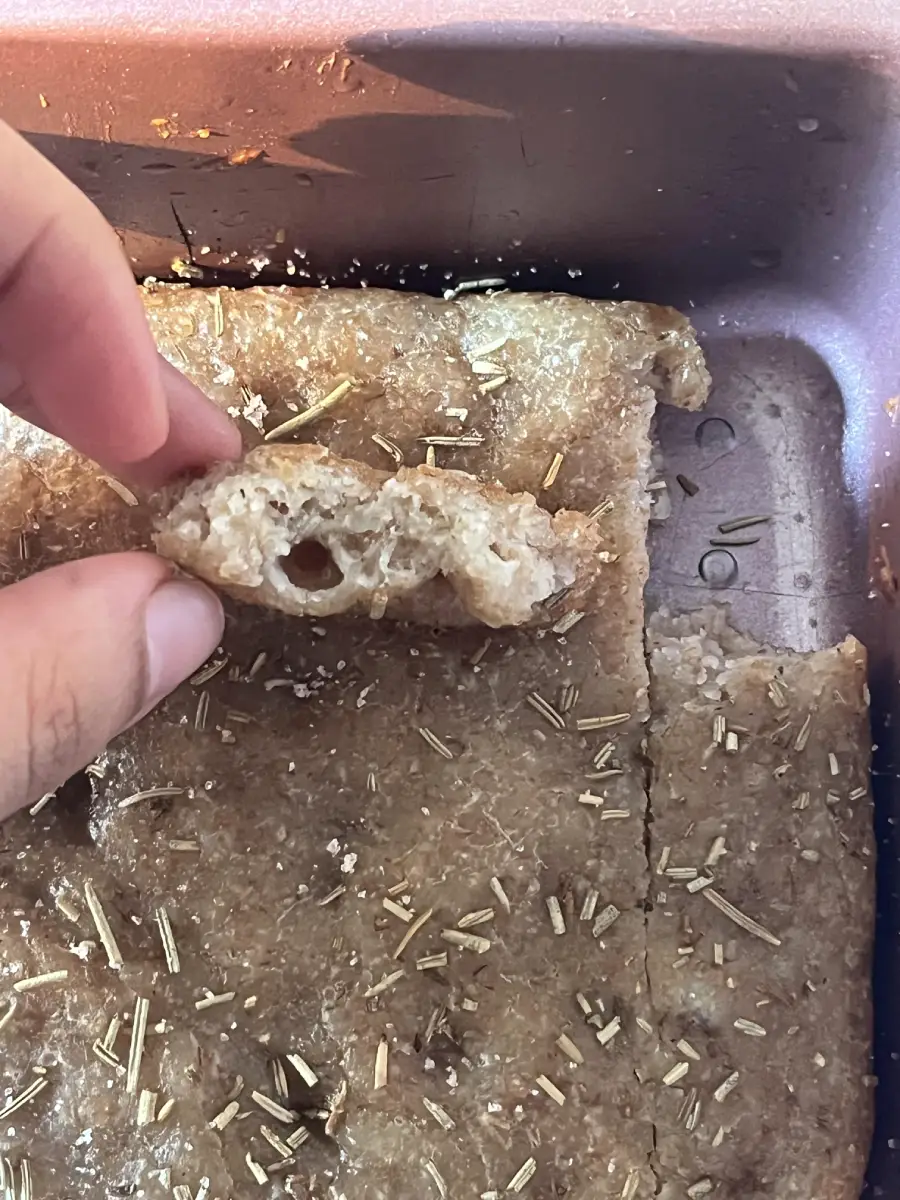 A hand holds up a slice of dense, homemade bread with a hole in the center. The bread is in a baking pan and topped with herbs, possibly rosemary.