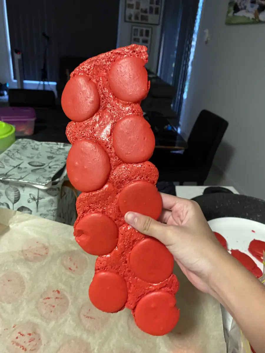 A hand holds a long, red, uneven sheet of failed macarons with round tops attached, in a kitchen setting.