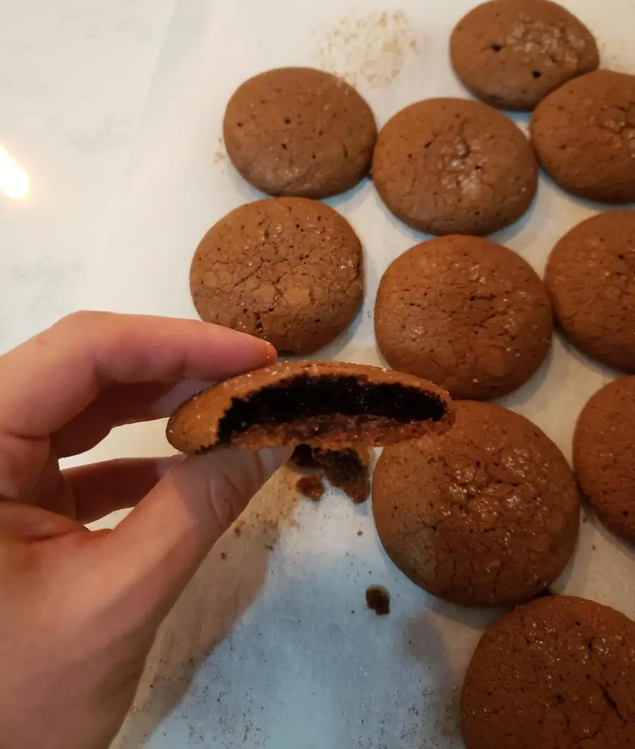 A hand holds a bitten chocolate cookie, showing a gooey center. More round, unbitten cookies sit on parchment paper in the background.
