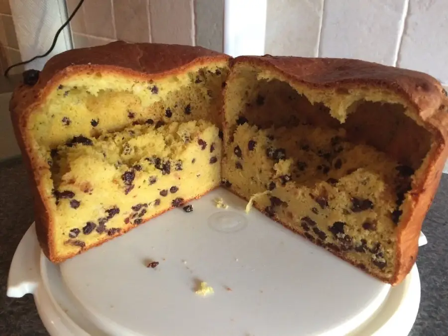 A round loaf of fruit cake, sliced in half to show a dense interior with scattered raisins or currants, sits on a white plate against a tiled kitchen backsplash.