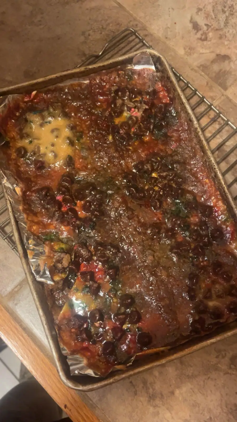 A metal baking tray lined with foil holds a dark, gooey, and unevenly baked dessert with visible chunks of colorful fruit or candy, and moist, sticky patches on top. The tray rests on a metal cooling rack.