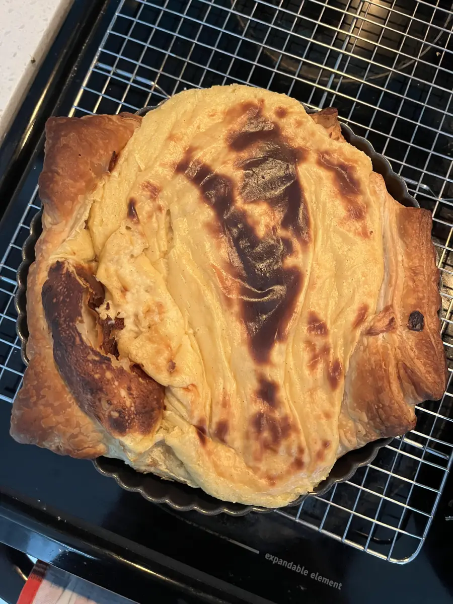 A slightly burnt, homemade pastry with a golden-brown top and uneven crust sits on a cooling rack over a baking tray.