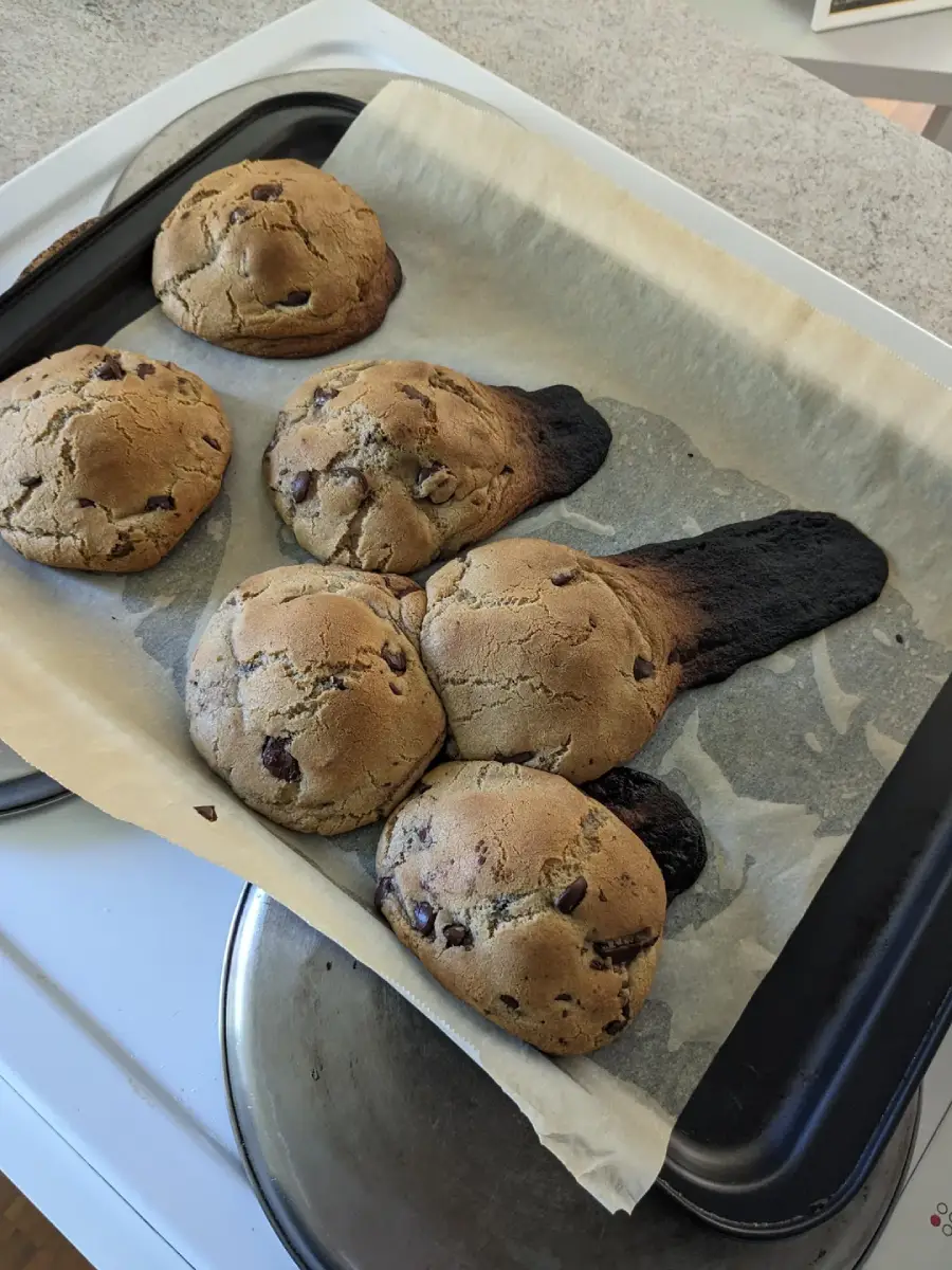Six chocolate chip cookies on a baking tray, with one cookie melted and burned, spreading a dark, charred streak onto the parchment paper beside the other round, baked cookies.