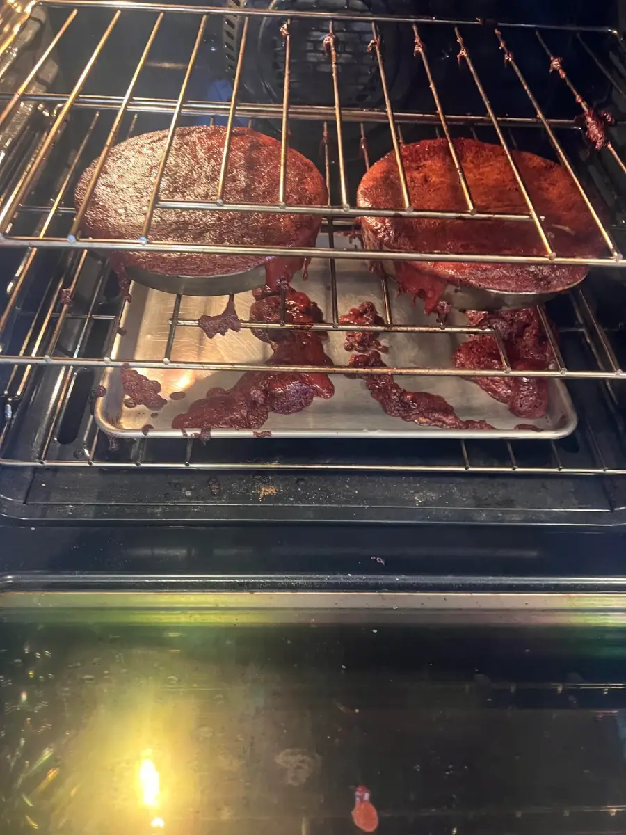 Two cake pans in an oven with batter overflowing and spilling onto the tray below, creating burnt, messy drips. The oven door is open, showing the baking mishap.