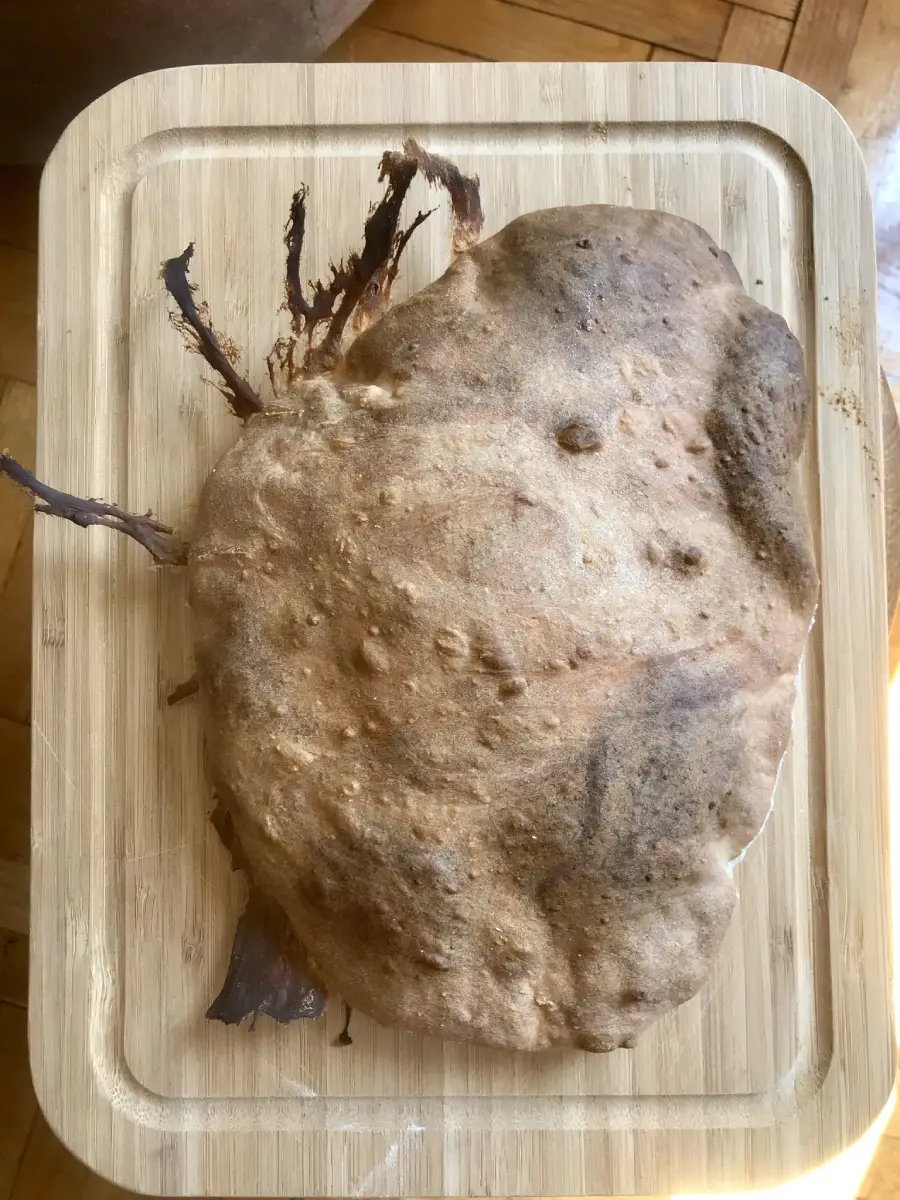 A misshapen, overcooked pizza with burnt edges sits on a wooden cutting board, displaying dark brown and unevenly baked crust.