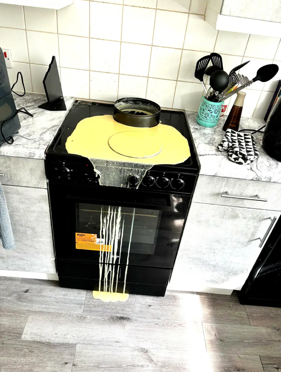 Cake batter has overflowed from a round pan in the oven, spilling onto the stovetop and dripping down the front of the oven onto the floor in a modern kitchen.