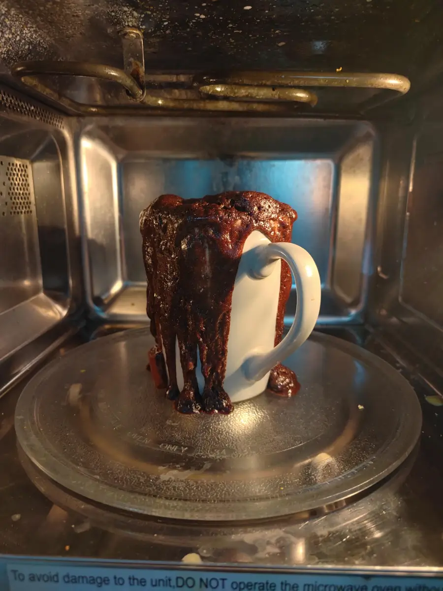 A white mug with chocolate cake overflowing and dripping down the sides sits on a glass microwave tray inside a microwave oven.
