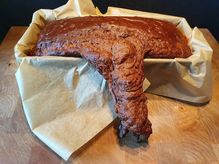 A loaf of bread has overflowed and spilled out of a parchment-lined baking tin, creating an irregular, lumpy shape that drapes over the edge onto a wooden surface.