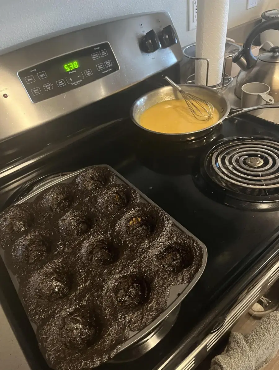 A stove with a muffin pan of baked chocolate muffins and a saucepan of yellow batter being whisked. The oven clock shows 5:38. Kitchen utensils and a paper towel roll are in the background.