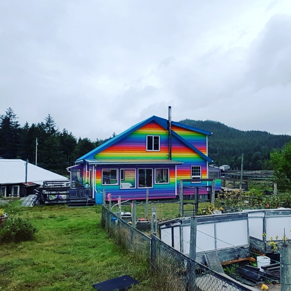A house painted with vibrant rainbow stripes stands in a grassy yard, surrounded by a fence and garden beds, with trees and hills in the background under a cloudy sky.