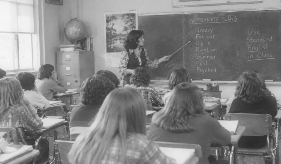 A teacher points to a chalkboard listing slang words in a classroom. Students sit at desks facing the teacher. The words “Use Standard English in Class” are written on the board.