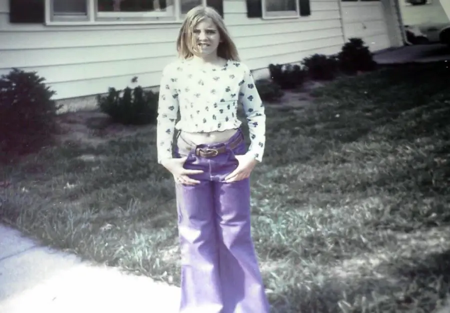 A young girl with long hair stands on grass near a sidewalk, wearing a long-sleeve floral top and high-waisted purple bell-bottom pants. A white house and shrubs are visible in the background.