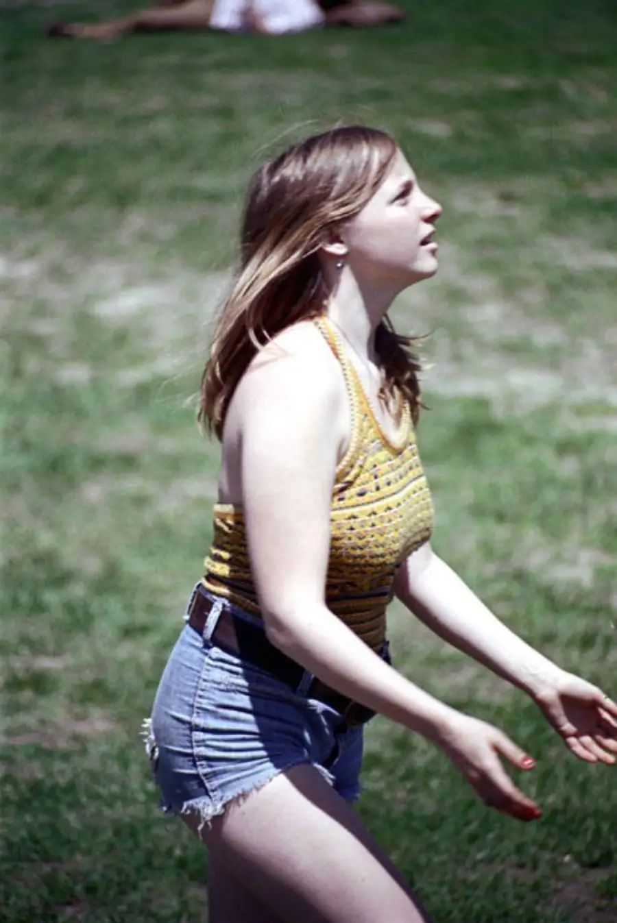 A young woman in a yellow patterned tank top and denim shorts stands outdoors on grass, looking upward with her arms slightly outstretched.