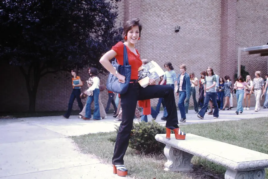 A woman in red stands on a bench outdoors, posing with one foot up. She wears platform shoes and holds books. Several people walk in the background near a brick building.