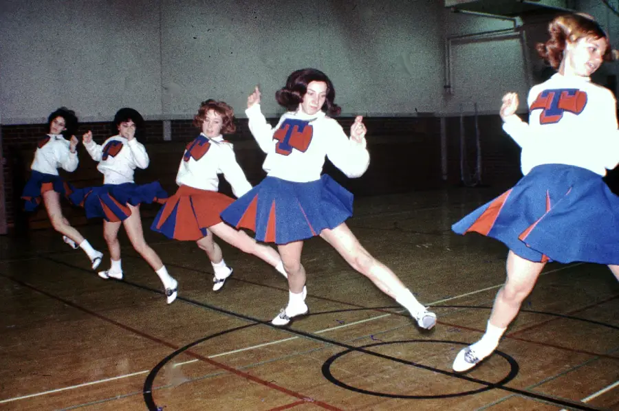 Five cheerleaders in matching blue and orange uniforms perform a synchronized kick routine in a gymnasium. A large “T” is displayed on the front of their sweaters.