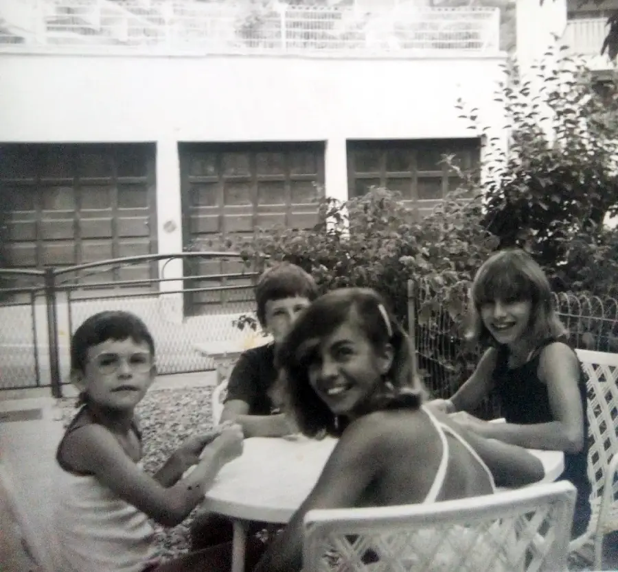 Four children sit around a round white table outdoors, smiling and looking at the camera. The photo is black and white, and behind them are bushes, a fence, and a building with garage doors.