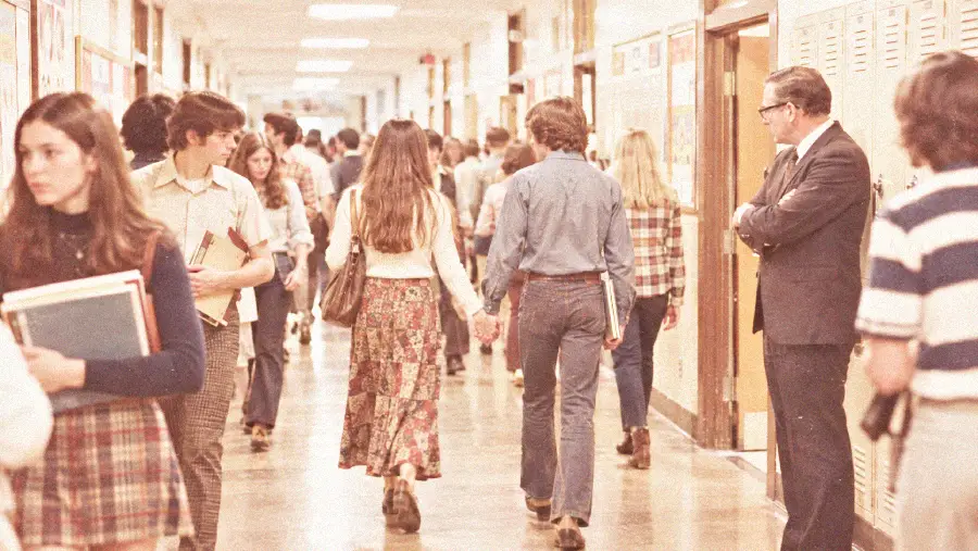 High school students walk through a crowded hallway. Two students in the center hold hands. Lockers line the walls, and an adult stands nearby observing. The scene appears to be from the 1970s.
