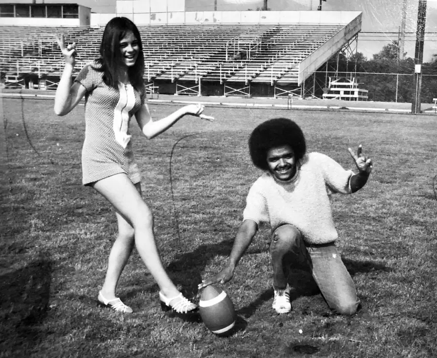 A woman stands and a man kneels on a football field holding a football upright. Both are smiling, making peace signs, and looking at the camera. Empty bleachers are visible in the background.