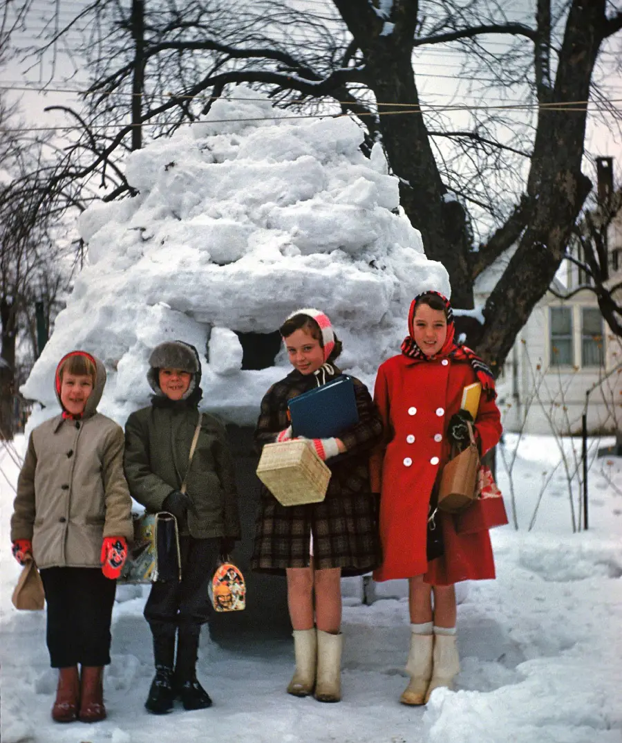Four children in winter coats and boots stand in snow in front of a large igloo made of snow, with leafless trees and a house in the background. Some children hold lunchboxes and bags.