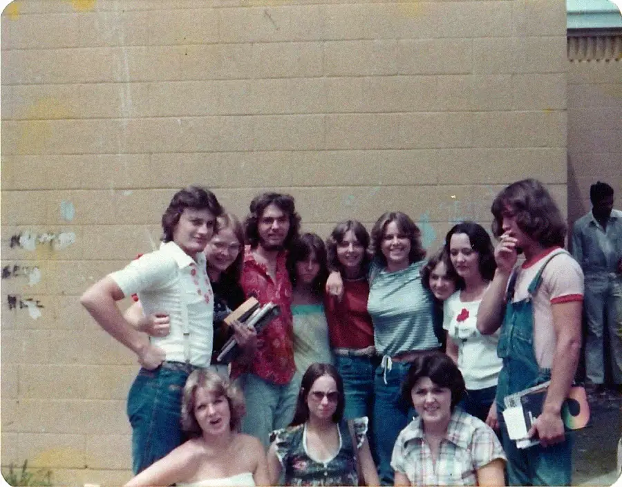 A group of teens, mostly smiling, pose together in front of a tan brick wall. Most wear casual 1970s clothes; some hold books. Two people sit in front. A man stands in the background on the right.