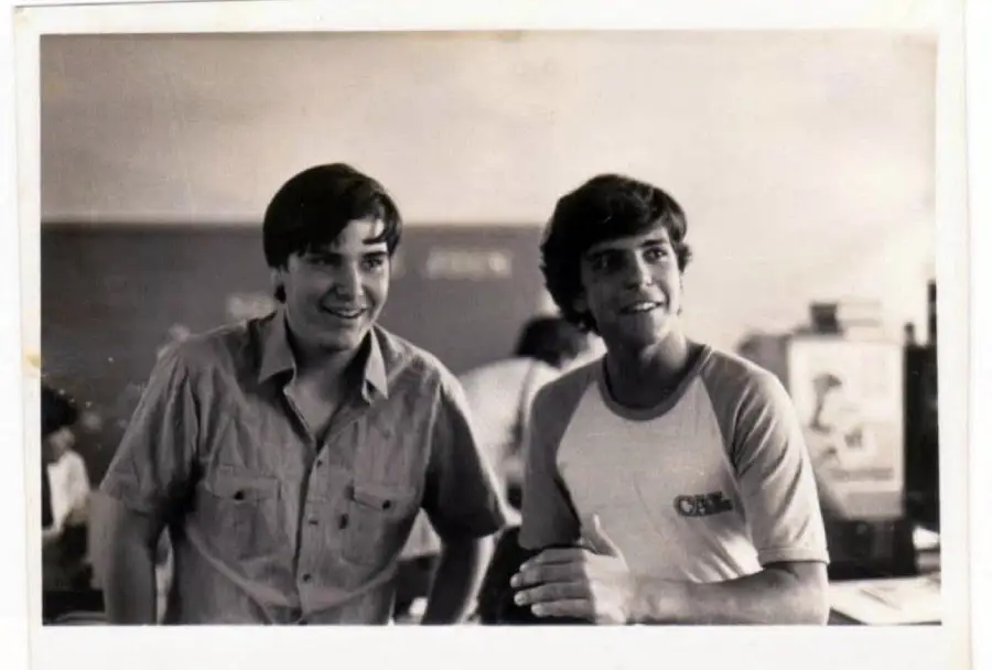 Two young men stand side by side indoors, smiling. One wears a button-up shirt, and the other wears a T-shirt. The background appears to be a classroom or casual setting. The photo is in black and white.