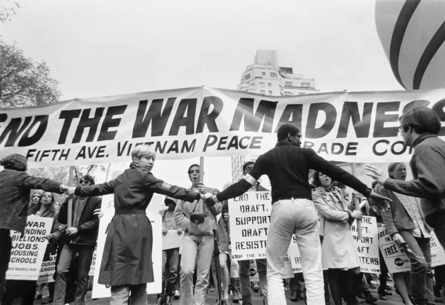 A group of protesters hold hands in front of a large banner reading “End the War Madness” during a Vietnam War protest, with signs and buildings visible in the background.
