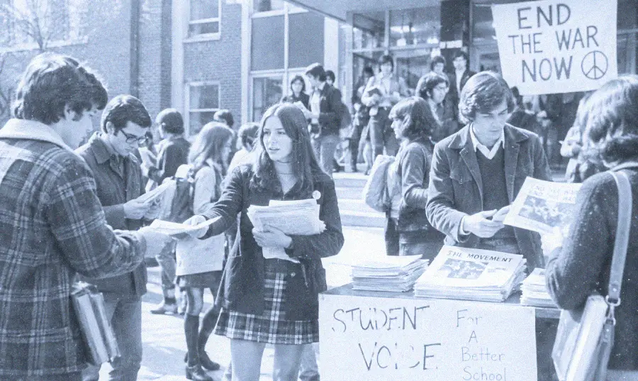 Students hand out newspapers and flyers at a campus event protesting war. A sign reads “END THE WAR NOW” and a table labeled “STUDENT VOICE” displays more papers. Several people gather, some holding protest signs.