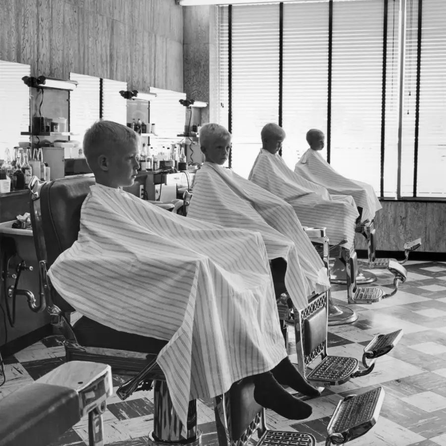 Three children sit in barber chairs, covered in striped capes, inside a vintage-style barbershop with large windows and mirrors along the wall.