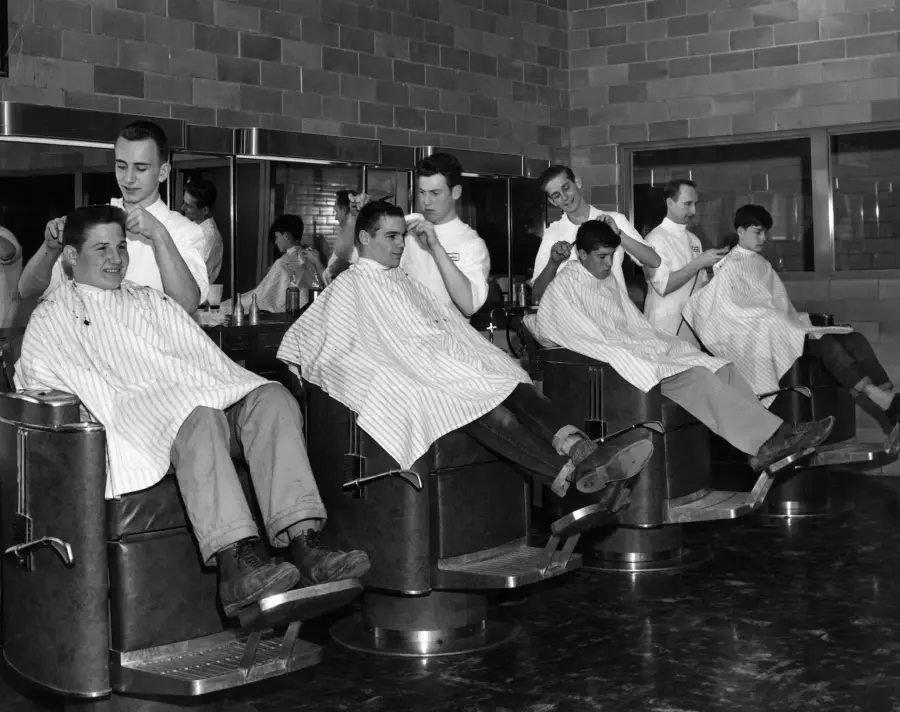 Six men sit in barber chairs getting haircuts from barbers in white coats. The barbershop has brick walls, mirrors, and a shiny floor. The scene appears busy and lively, with everyone focused on their tasks.
