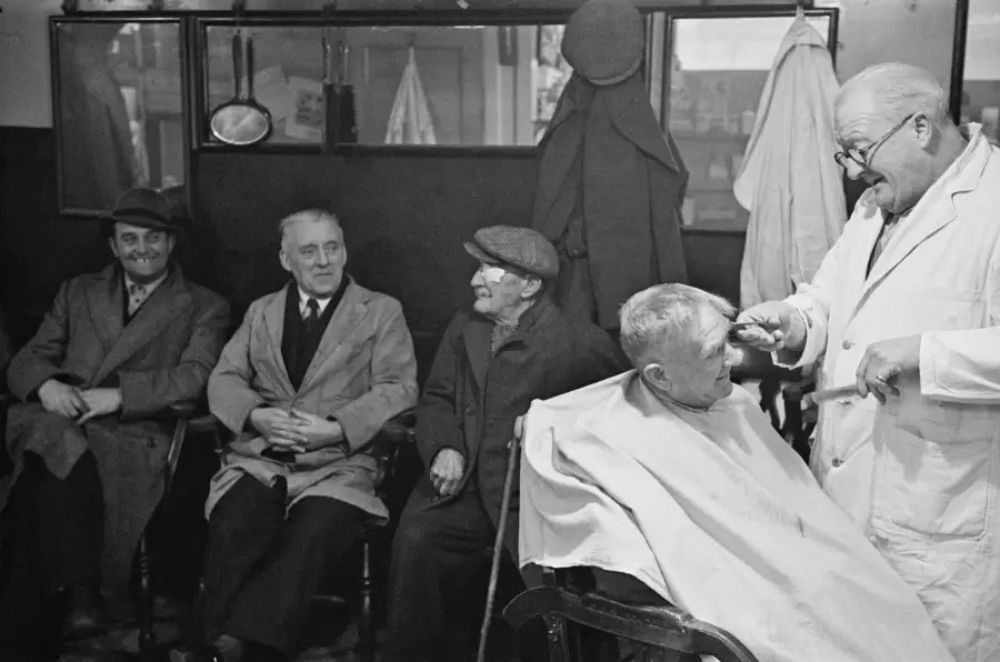 Four older men sit waiting in a barbershop while a barber in a white coat cuts a young boy’s hair. Coats and hats hang on the wall behind them. The scene appears relaxed and social.