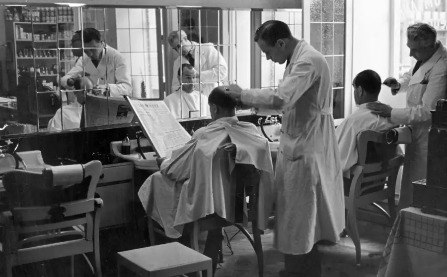 Black and white photo of three barbers cutting hair for three men seated in chairs, one man reads a newspaper. Mirrors line the wall, reflecting the barbers and clients. Shelves of products are visible in the background.