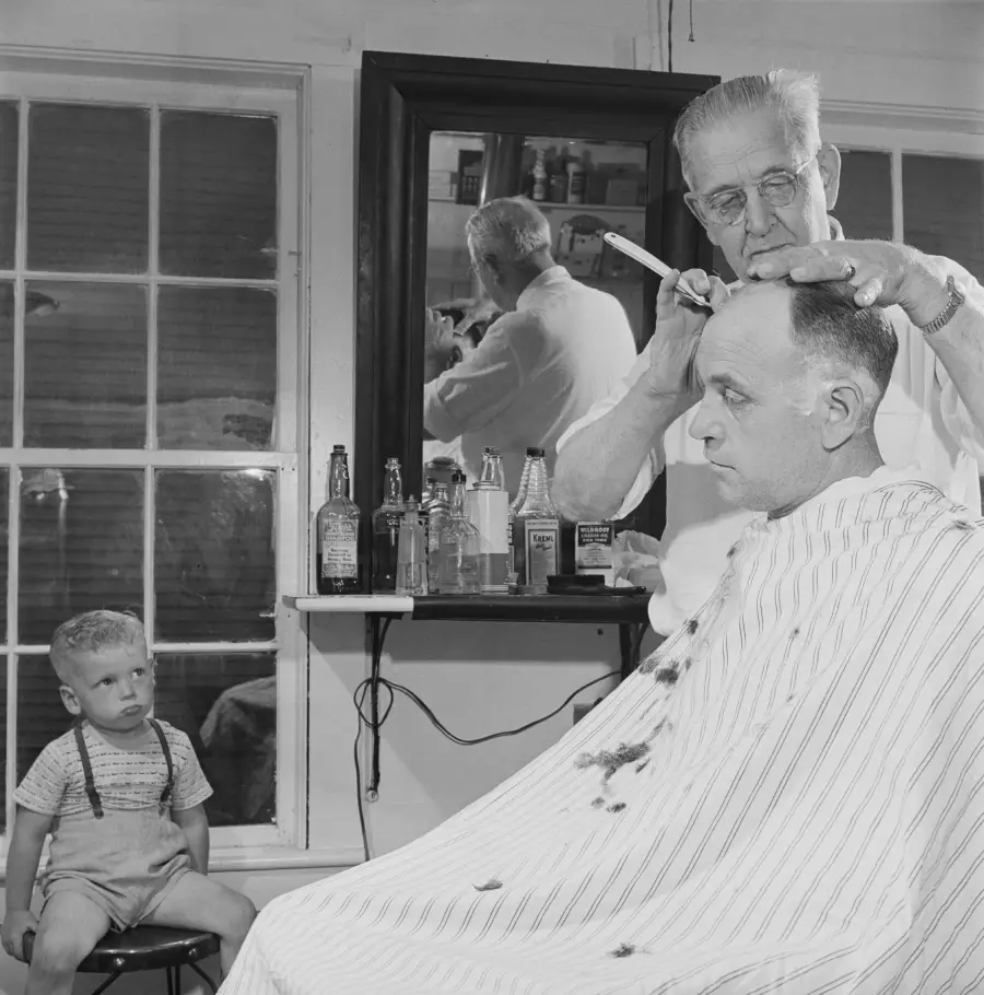 A barber cuts a man’s hair in a barbershop while a young boy in suspenders sits nearby, watching. Bottles sit on a shelf below a mirror reflecting the scene.